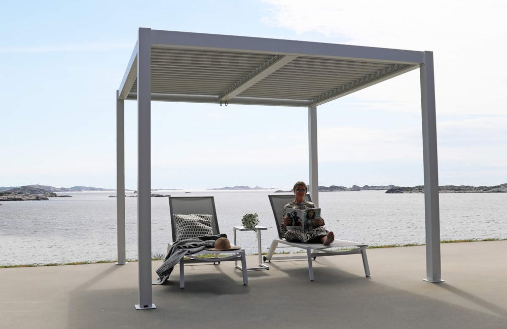 Stavern pergola_10 Hvit pergola med to solsenger under og dame som leser blad ved sjøen