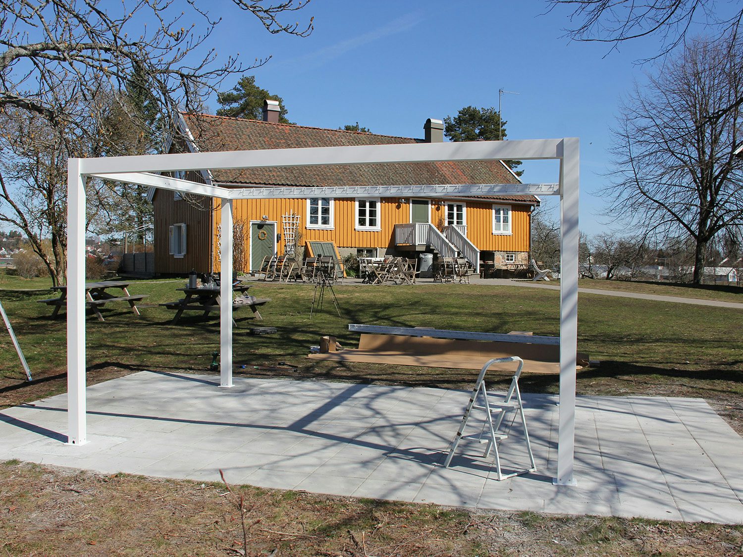 Pergola uten lameller foran gult hus. Foto